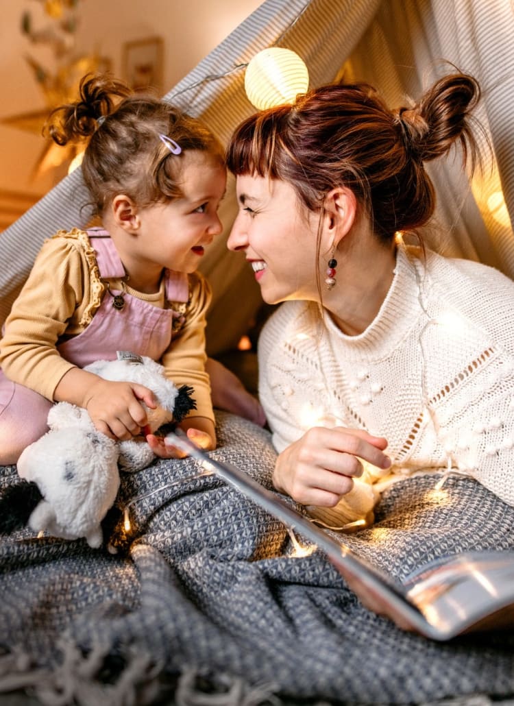 Mother reading a book to her toddler daughter in a dimly lit tent in child's bedroom.